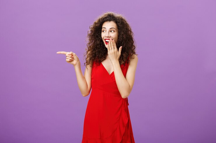 Photo gratuit une fille rit devant un mec maladroit qui fait une erreur lors d'une fête couvrant le sourire avec un regard de paume et pointant vers la gauche tout en bavardant avec des copines se tenant joyeusement dans une élégante robe rouge sur un mur violet.