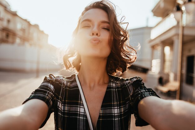 Fille romantique faisant selfie en journée chaude et ensoleillée. Adorable femme européenne debout dans la rue avec l'expression du visage qui s'embrasse.