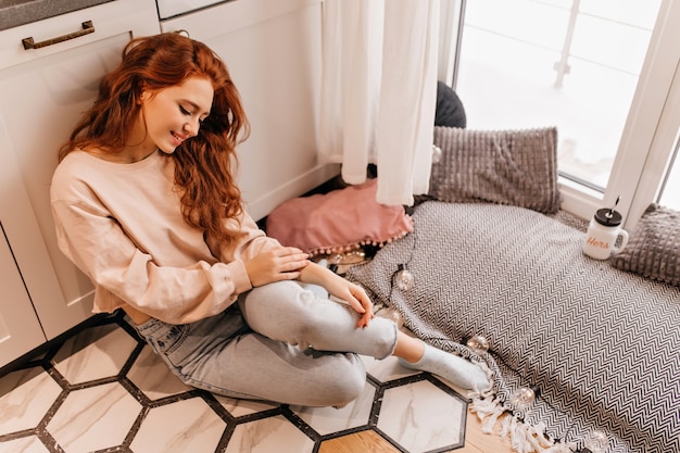 Fille romantique avec une longue coiffure assise sur le sol. Ginger jolie dame passant le week-end à la maison.