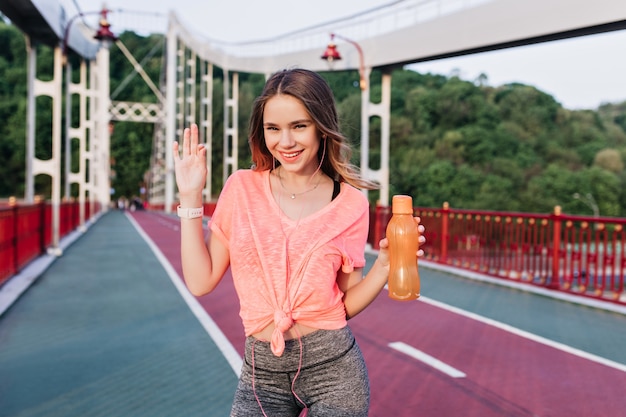 Fille romantique en t-short rose s'amusant sur le stade. Adorable jeune femme avec une bouteille d'eau en riant de la piste de cendre.