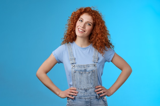 Une Fille Rousse Aux Cheveux Bouclés Et Séduisante, Motivée Et Gaie, Se Tient La Main à La Taille, Pose Confiante, Sa Propre Tête Inclinable, Souriante, Ravie, Sûre De Soi, Inspirée, Crée.