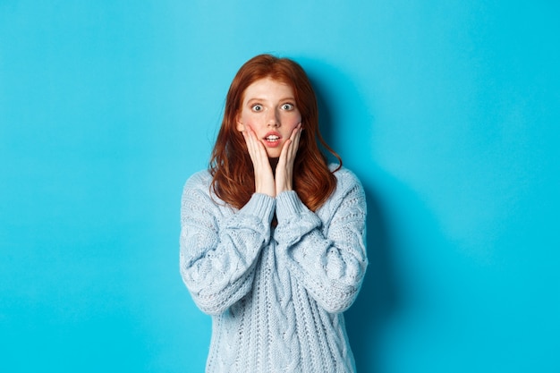 Une fille rousse choquée regardant la caméra sans voix, exprime son incrédulité et son étonnement, debout en pull sur fond bleu.