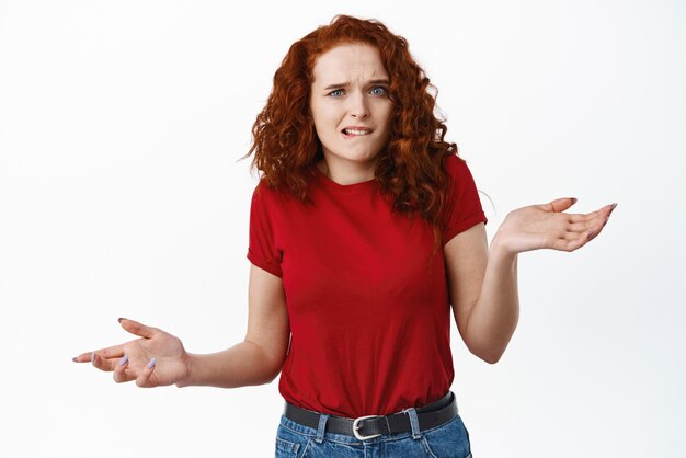 Fille rousse confuse et perplexe en t-shirt haussant les épaules et se mordant la lèvre se sentant mal à l'aise de ne pas savoir ne peut pas comprendre quelque chose debout désemparé sur fond blanc