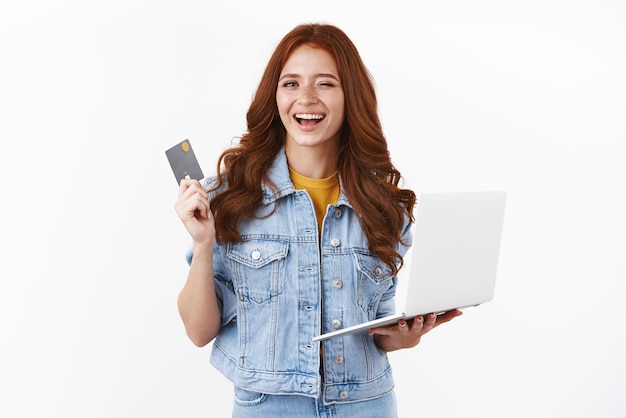 Fille rousse détendue et insouciante assurant que tout va bien tant qu'elle a un dépôt montrant une carte de crédit noire clin d'œil effronté tenir un ordinateur portable souriant satisfait en offrant un cadeau pour un ami dans un magasin Internet