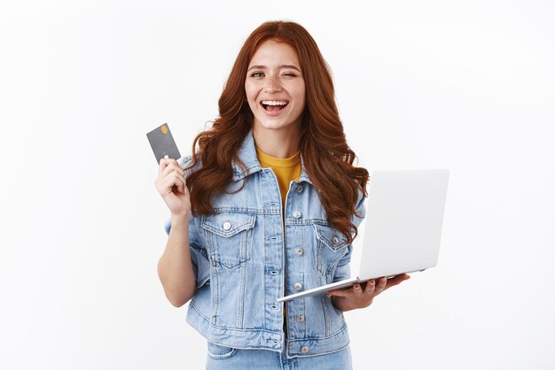 Fille rousse détendue et insouciante assurant que tout va bien tant qu'elle a un dépôt montrant une carte de crédit noire clin d'œil effronté tenir un ordinateur portable souriant satisfait en offrant un cadeau pour un ami dans un magasin Internet