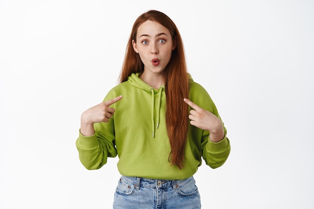 Fille rousse excitée et surprise se pointant du doigt, l'air amusée, choisie ou gagnante, auto-promotion, veut participer à l'événement, fond blanc