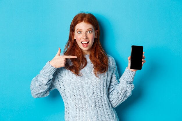 Fille rousse impressionnée pointant sur l'écran du téléphone, montrant une application pour smartphone ou une offre en ligne et souriante excitée, debout en pull sur fond bleu.