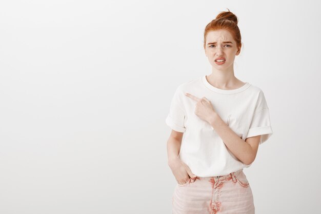 Fille rousse indécise et incertaine fronçant les sourcils perplexe et pointant vers la gauche