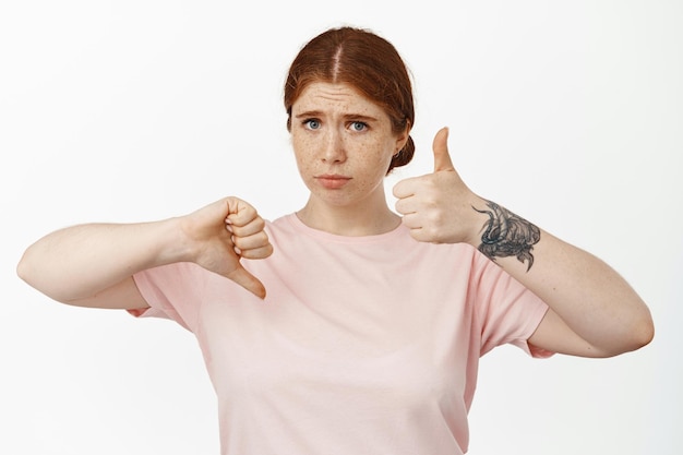 Fille rousse indécise montrant les pouces vers le haut, le pouce vers le bas et l'air perplexe, jugeant le résultat moyen pas mauvais, hésitant à évaluer quelque chose, debout sur fond blanc