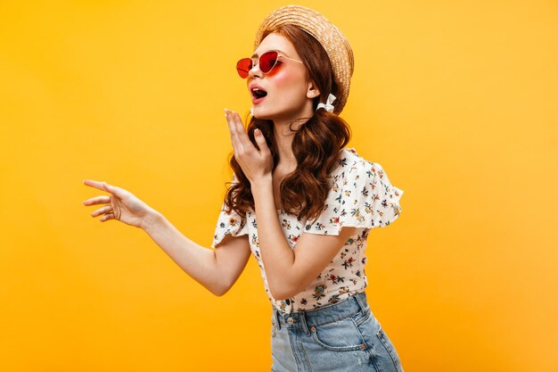 Fille rousse à lunettes rouges et chapeau de paille souffle baiser Portrait de femme en blouse avec des fleurs sur fond orange