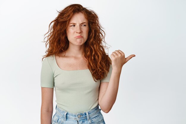 Fille rousse mécontente boudant en fronçant les sourcils avec aversion pointant et regardant le côté droit bouleversé debout en t-shirt décontracté sur fond blanc