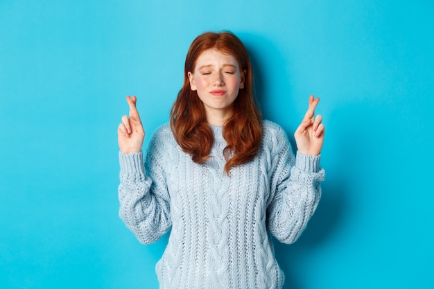 Une fille rousse pleine d'espoir fait un vœu, croise les doigts pour la bonne chance, sourit et anticipe une bonne nouvelle ou un résultat positif, debout sur fond bleu.