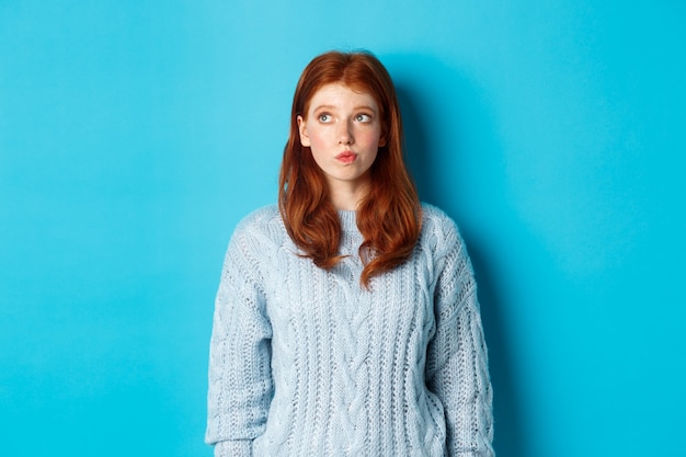 Fille Rousse Rêveuse Pensant Ou Prenant Une Décision, Regardant Le Logo Du Coin Supérieur Gauche, Debout Sur Fond Bleu.