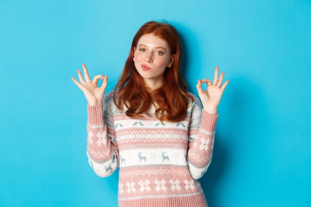 Une fille rousse satisfaite et fière hoche la tête en signe d'approbation, montrant un signe d'accord, pas mal ou un geste de louange, debout sur fond bleu