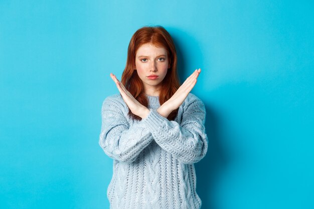 Une fille rousse sérieuse à l'air confiante, montrant un geste croisé pour arrêter et interdire l'action, debout sur fond bleu.