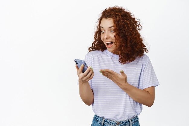 Fille rousse surprise et excitée lisant de bonnes nouvelles, heureuse du message de son téléphone portable, réagissant joyeusement à la notification sur smartphone sur blanc.