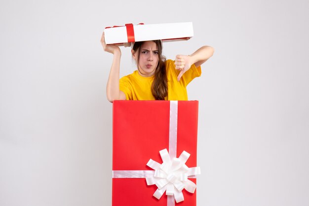 Fille sans joie avec bonnet de Noel faisant le pouce vers le bas signe debout derrière un grand cadeau de Noël sur blanc