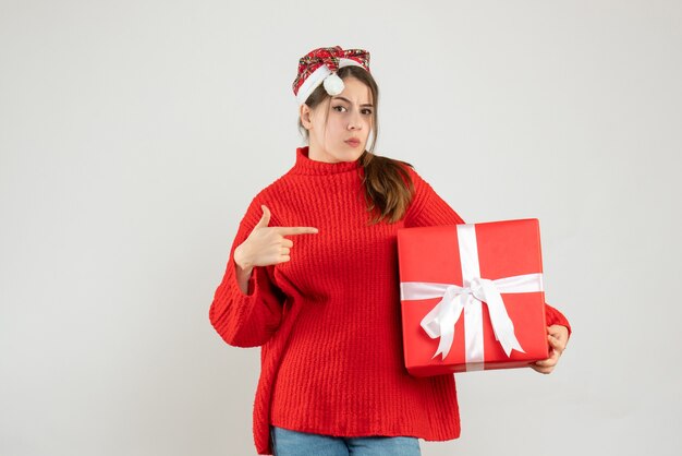 Fille sceptique avec Bonnet de Noel tenant présent pointant avec le doigt la boîte quelque chose debout sur blanc