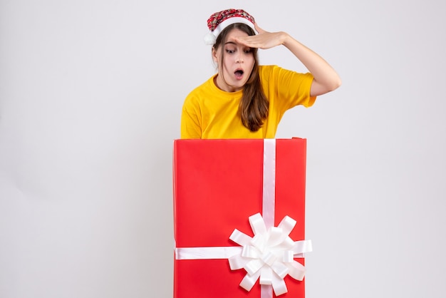 Fille se demande avec bonnet de Noel debout derrière un grand cadeau de Noël sur blanc