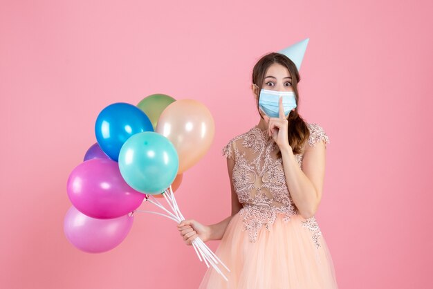 Fille se demande avec chapeau de fête et masque médical faisant signe chut tenant des ballons colorés sur rose