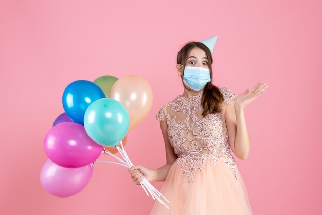 fille se demande avec chapeau de fête et masque médical tenant des ballons colorés sur rose