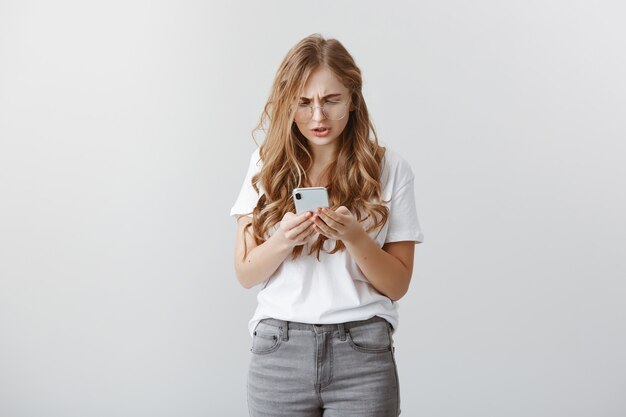 Fille se sentant mal à l'aise d'envoyer un message au mauvais numéro. Portrait de jolie jeune femme troublée dans des verres, jurant tout en fronçant les sourcils et à la dérangée à l'écran du smartphone, navigation en réseau