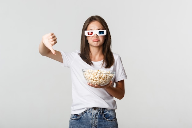 Fille séduisante déçue dans des lunettes 3D, tenant du pop-corn et montrant les pouces vers le bas, n'aime pas le film.