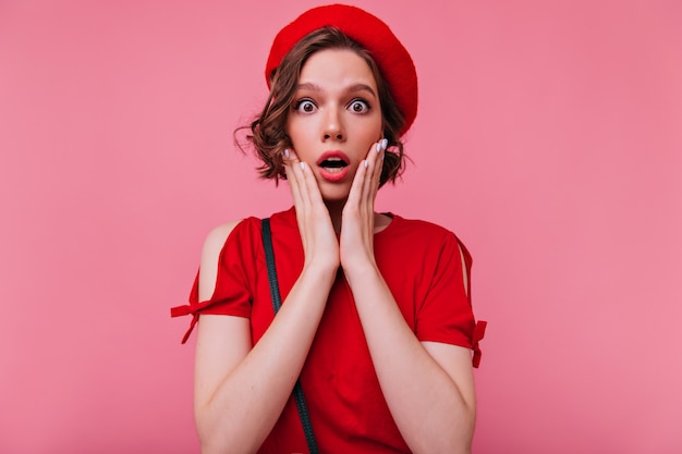 Fille séduisante étonnée avec des cheveux bruns ondulés à la recherche. Portrait intérieur de l'adorable dame française exprimant des émotions surprises.