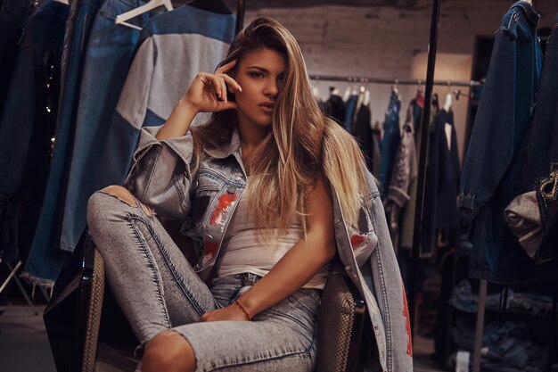 Fille séduisante portant un jean en détresse et une veste en jean regardant une caméra tout en étant assise sur une chaise dans la cabine d'essayage d'un magasin de vêtements.