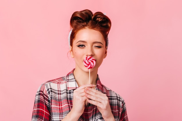 Fille sensuelle au gingembre mangeant des bonbons rouges. Vue de face de l'élégante femme pin-up avec sucette isolée sur l'espace rose.