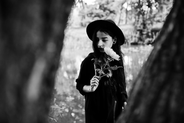 Fille sensuelle tout en lèvres rouges noires et chapeau Goth dramatique femme tient une fleur de chrysanthème blanche à portée de main contre le lac silencieux Portrait noir et blanc