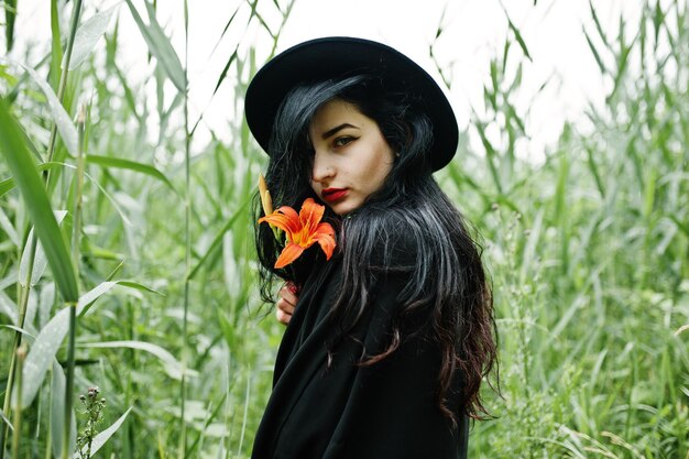 Fille sensuelle tout en lèvres rouges noires et chapeau Goth dramatique femme tient une fleur de lys orange sur un roseau commun