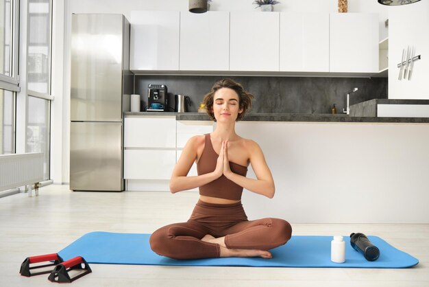 Fille souriante, athlète, sportive, faisant du yoga à la maison en vêtements de sport, assise sur un tapis de yoga en caoutchouc