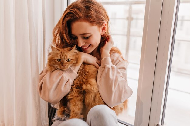 Fille souriante détendue jouant avec son chat moelleux. Plan intérieur d'une femme incroyable tenant un animal de compagnie.