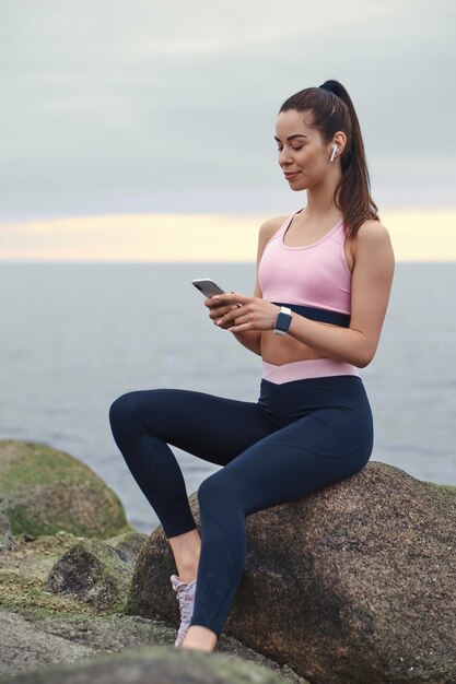 Une fille souriante et heureuse discute par téléphone portable tout en étant assise sur la pierre au bord de la mer calme.