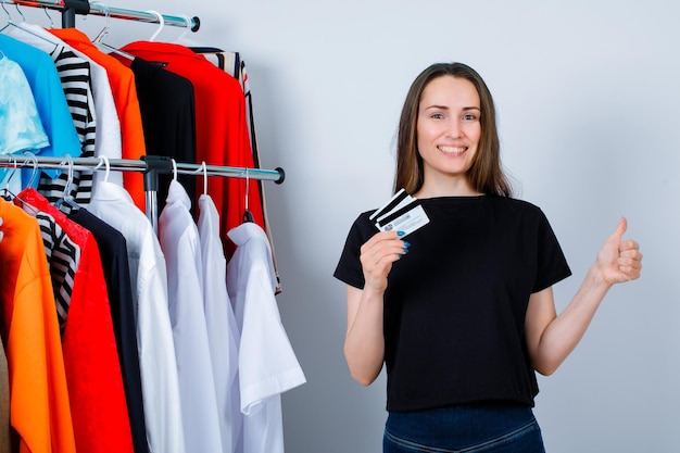 Une fille souriante montre un geste parfait en tenant des cartes de crédit sur fond de vêtements