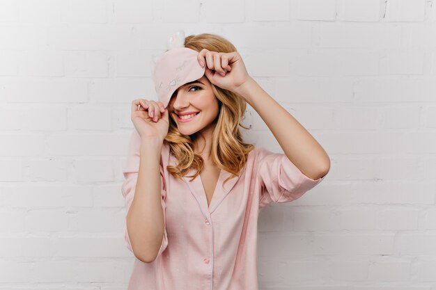 Fille souriante pâle aux cheveux bouclés posant de manière ludique sur un mur blanc. Femme effrayante en masque pour les yeux et pyjama en soie riant à la maison.