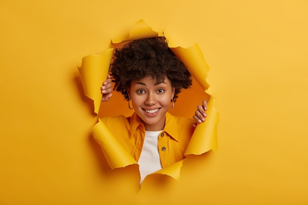 Fille souriante positive avec coupe de cheveux bouclés, vêtue d'une veste jaune à la mode, pose à travers un fond de papier déchiré, montre des dents blanches, profite d'une bonne journée, de bonne humeur.