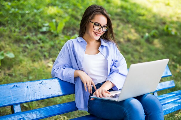 Fille souriante en t-shirt bleu s'asseoir sur le banc dans le parc et utiliser son nouvel ordinateur portable