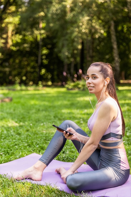 Fille sportive assise sur un tapis de yoga écoute une musique dans le parc