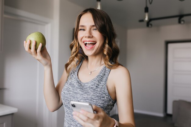 Fille sportive exprimant le bonheur tout en posant avec des fruits. Tir intérieur d'une femme bouclée détendue avec apple et smartphone.