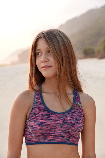 Fille sportive vêtue d'un haut décontracté, a un corps de remise en forme sain, se sent rafraîchie après l'entraînement en plein air, regarde pensivement de côté, pose à l'extérieur