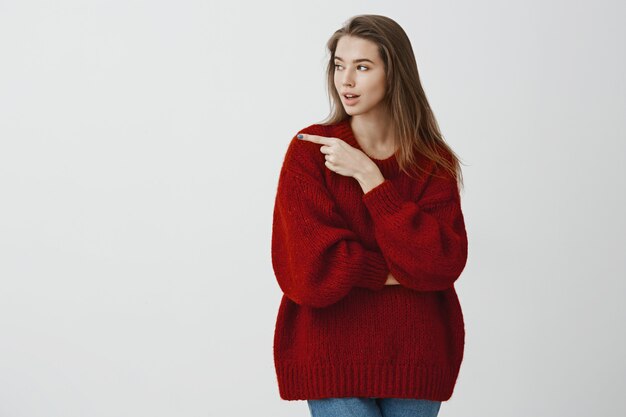 Fille suggérant des amis pour frapper la barre dans cette direction. Concentré de jolie femme sérieuse en pull rouge lâche, regardant et pointant vers la gauche, discutant d'un café intéressant, debout sur un mur gris