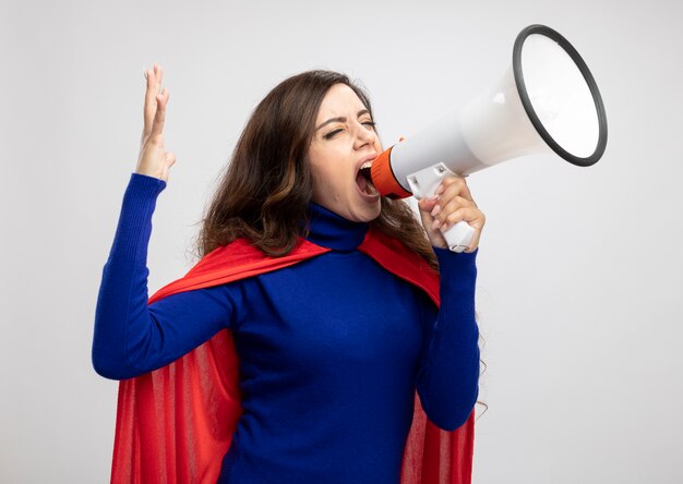 Fille de super-héros caucasien agacé avec cape rouge se dresse avec la main levée et crie dans haut-parleur isolé sur mur blanc avec espace copie