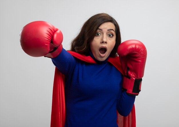 Fille de super-héros caucasien choqué avec cape rouge portant des gants de boxe fait semblant de frapper