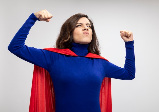 Photo gratuite fille de super-héros caucasien furieux avec cape rouge tend les biceps et regarde à côté sur blanc