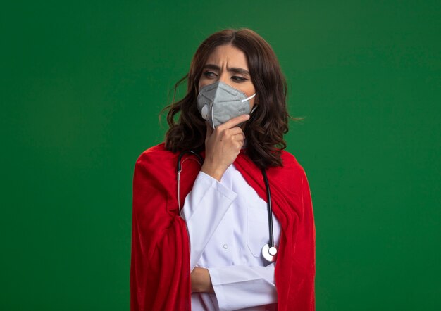 Fille de super-héros caucasien réfléchie en uniforme de médecin avec cape rouge et stéthoscope portant un masque médical