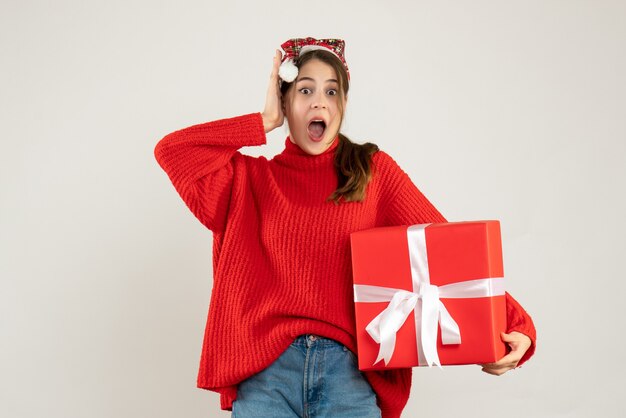 fille surprise avec bonnet de noel tenant présent mettant la main à sa tête debout sur blanc