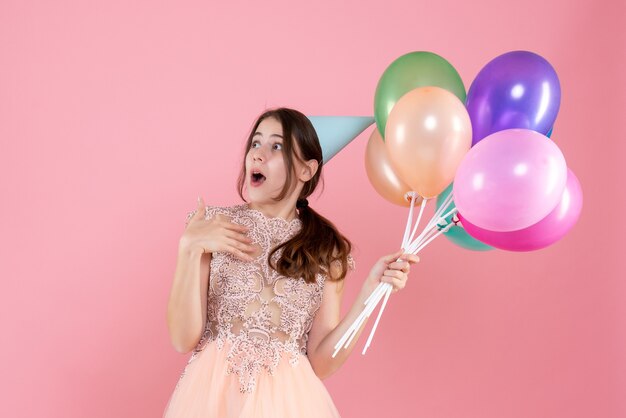 fille surprise avec chapeau de fête tenant des ballons en regardant quelque chose sur le rose