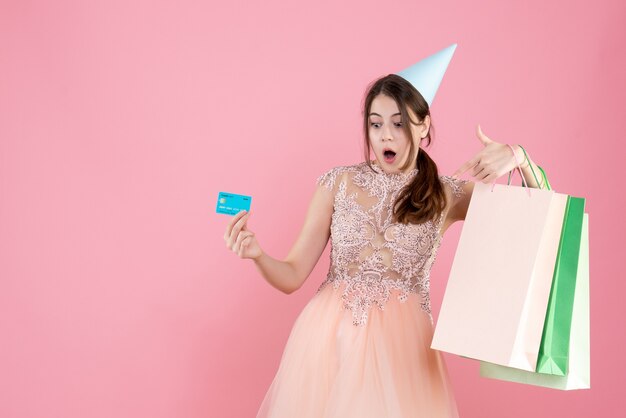 fille surprise avec chapeau de fête tenant une carte de crédit et des sacs à provisions sur rose